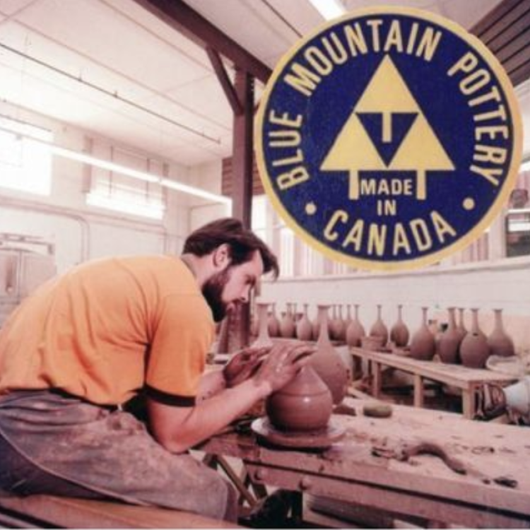 White man in orange t-shirt with smock covering pants. The man is shaping the clay pot in a pottery studio. There are clay pots lining the back wall on a wood table. In the top right corner there is a superimposed Blue Mountain Pottery Logo. The circular logo has a dark blue back with Blue Mountain Pottery text in beige around the top perimeter of the logo, three beige triangles in a pyramid shape with lines out the bottom representing tress in the middle, along the bottom reads Made In Canada 