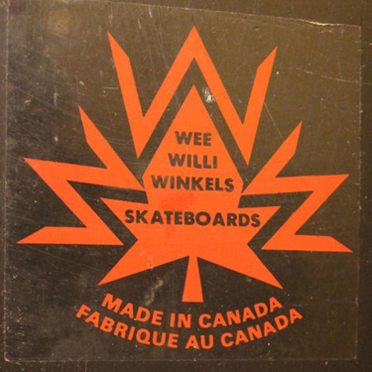 A Wee Willi Winkels Skateboards logo with a brown background. The logo is orange and shaped like a maple leaf with three letter W's on the perimeter creating the points of the leaf. In the middle of the leaf is brown text reading Wee Willi Winkels Skateboards. Below the leaf is orange text reading Made in Canada Fabrique au Canada.