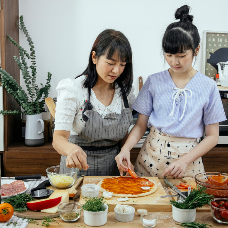 Two women wearing aprons assembling a pizza on the counter in front of them, various ingredients spread out around them.