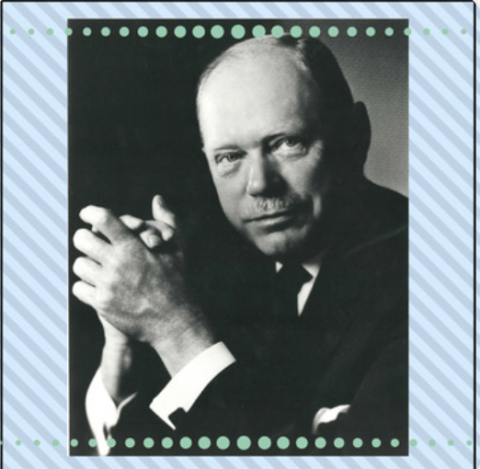 A white man clasping his hands as he looks forward. The man is wearing a dark suit and has some facial hair.