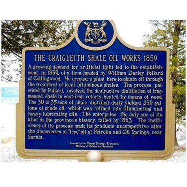 A photograph of a blue commemorative plaque with a gold border and text situated outdoors. The text details the history of The Craigleith Shale Oil Works in 1859. In the background behind the plaque is a snow covered ground and evergreen trees.