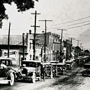 A black and white photograph of a 1920's street with a row of buildings and cars parked along the left side of the road. In the foreground are two people, one sitting in the driver's seat of a car and one man standing outside of a car on the driver's side. In the midground, a car can be seen driving on the road.