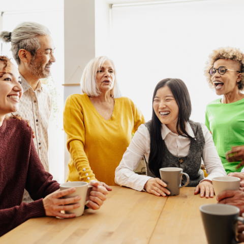 a group of people gather around a wood table, some holding coffee, smiling, and others standing and talking.
