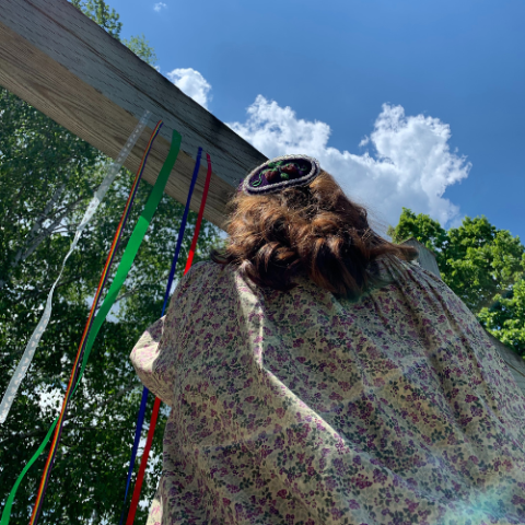 Woman in Indigenous Dress and hair accessory ties ribbons on a wood beam, image is viewed from the floor looking towards the sky