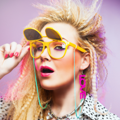 Woman dressed in 1980s attire, crimped blonde hair, yellow glasses with green chain, pink "love" earring, and bright pink lipstick.