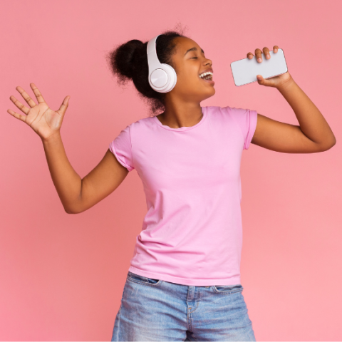 Woman with headphones on holding up phone and singing into the end of it.