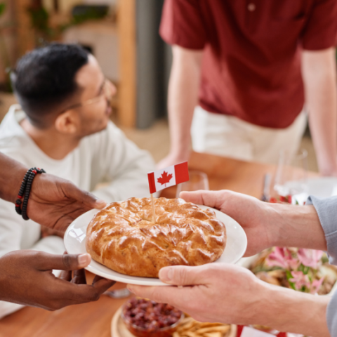 One person handing another person a plate with a pie on it that has a Canada flag stuck in the middle