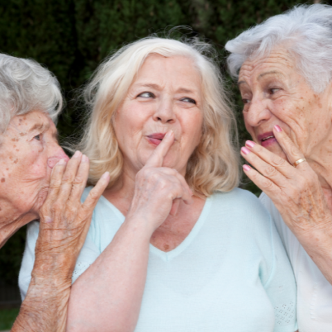 Three elderly women standing side by side. Women in middle is holding finger up to mouth in "shh" motion. Two women at her sides are whispering in her ears.