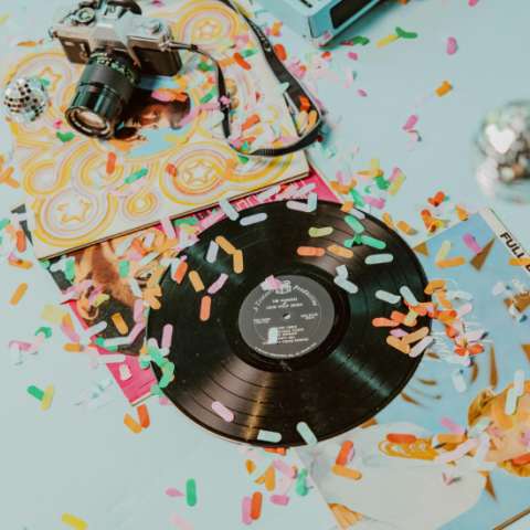 Vinyl records and an old fashioned camera sitting on blue background with confetti surrounding. 